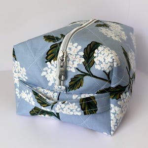 Puede incluir: Un neceser acolchado azul claro con un estampado floral de flores blancas y hojas verdes. Tiene un cierre de cremallera plateada y un asa pequeña. La tela está acolchada con hilo blanco.