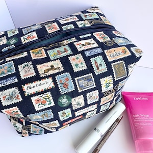May include: A navy blue zippered pouch with a colorful postage stamp print. The pouch is made of fabric and has a zipper closure. The pouch is lying on a white surface.