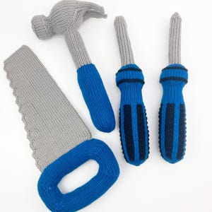 May include: A collection of knitted toy tools, including a hammer, two screwdrivers, and a saw. The hammer and screwdrivers have blue handles, while the saw and hammer heads are gray. The tools are arranged on a white surface.