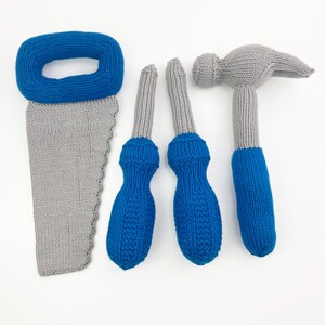 May include: A collection of knitted toy tools, including a gray saw with a blue handle, two blue and gray screwdrivers, and a gray hammer with a blue handle. The toys are designed for children and are made of soft, textured yarn.