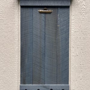 May include: A dark gray wooden wall organizer featuring a vertical plank design with a small top shelf, a brass-toned metal clip at the top center, and four black metal hooks attached to a horizontal base piece at the bottom.