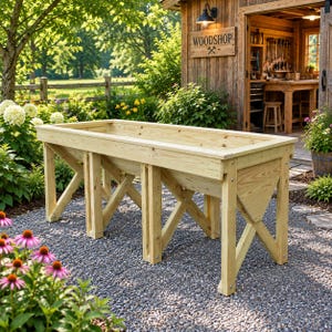 Puede incluir: Un bancal elevado rectangular construido con madera natural de color claro. La jardinera presenta una estructura robusta con patas en forma de cruz y un diseño de cubeta profunda abierta, situada al aire libre sobre grava.