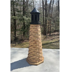 May include: A tall, decorative lighthouse with a black top and a light brown, brick-like structure. The lighthouse is made of wood and stands on a concrete surface, with a forest in the background.