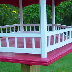 Peut inclure: Un mangeoire &agrave; oiseaux rouge et blanche avec une plateforme hexagonale et une balustrade de cl&ocirc;ture en bois blanc. La mangeoire est mont&eacute;e sur un poteau en bois.