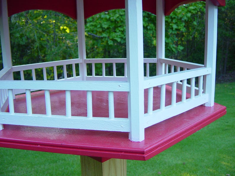 Peut inclure: Un mangeoire &agrave; oiseaux rouge et blanche avec une plateforme hexagonale et une balustrade de cl&ocirc;ture en bois blanc. La mangeoire est mont&eacute;e sur un poteau en bois.