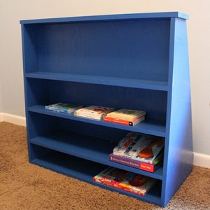 May include: A blue wooden bookshelf with four shelves, filled with children's books. The bookshelf is angled and has a triangular shape on the right side.