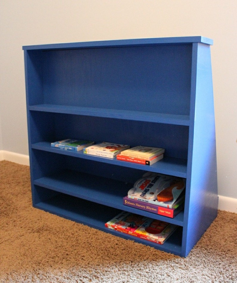 May include: A blue wooden bookshelf with four shelves, filled with children's books. The bookshelf is angled and has a triangular shape on the right side.
