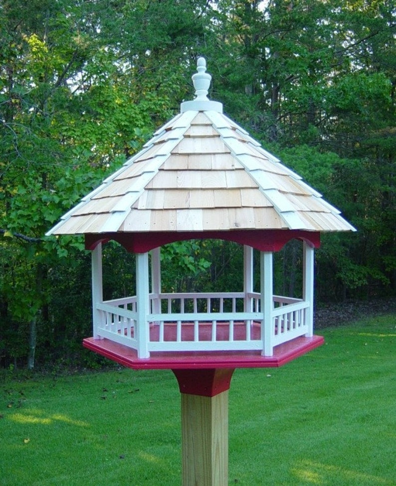 Peut inclure: Un mangeoire &agrave; oiseaux blanche et rouge avec un toit en bardeaux et un fleuron d&eacute;coratif. La mangeoire a une base hexagonale et une balustrade blanche autour du sommet.