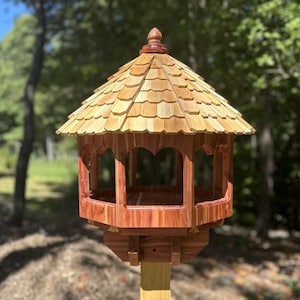 May include: A wooden bird feeder with a cedar roof and a hexagonal base. The feeder has a decorative finial on top and is mounted on a wooden pole.
