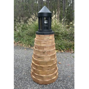May include: A wooden lighthouse with a black top and a series of stacked, octagonal wooden sections. The top section is black with a glass enclosure. The lighthouse is outdoors, set against a backdrop of trees and foliage.