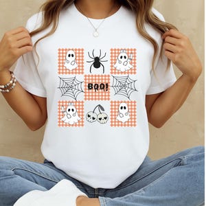 May include: A white t-shirt featuring a grid pattern of orange and white checkered squares. The design includes illustrations of ghosts with stethoscopes, a black spider, spiderwebs, a pair of skull cherries, and the text BOO in black letters.