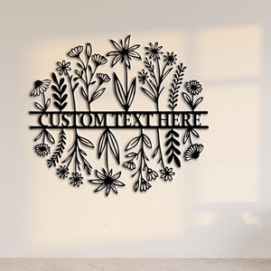 May include: Black metal floral wall art with the text "CUSTOM TEXT HERE" in a circular design. The artwork features various stylized flowers and foliage. The piece is mounted on a white wall, with a wooden chair and window in the background.