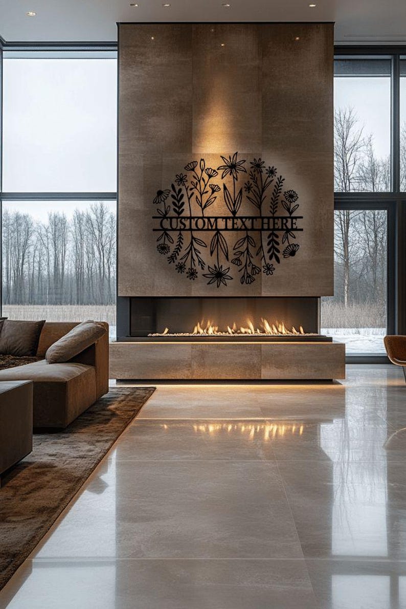 May include: A contemporary living room featuring a fireplace. A black metal floral design with the text "CUSTOM TEXT HERE" is mounted on the wall above the fireplace. The room has large windows with a view of trees.