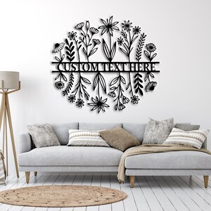 May include: Black metal floral wall art in a circular design. The artwork features stylized flowers and foliage around a horizontal bar with the text "CUSTOM TEXT HERE". It is mounted on a white wall above a grey sofa.