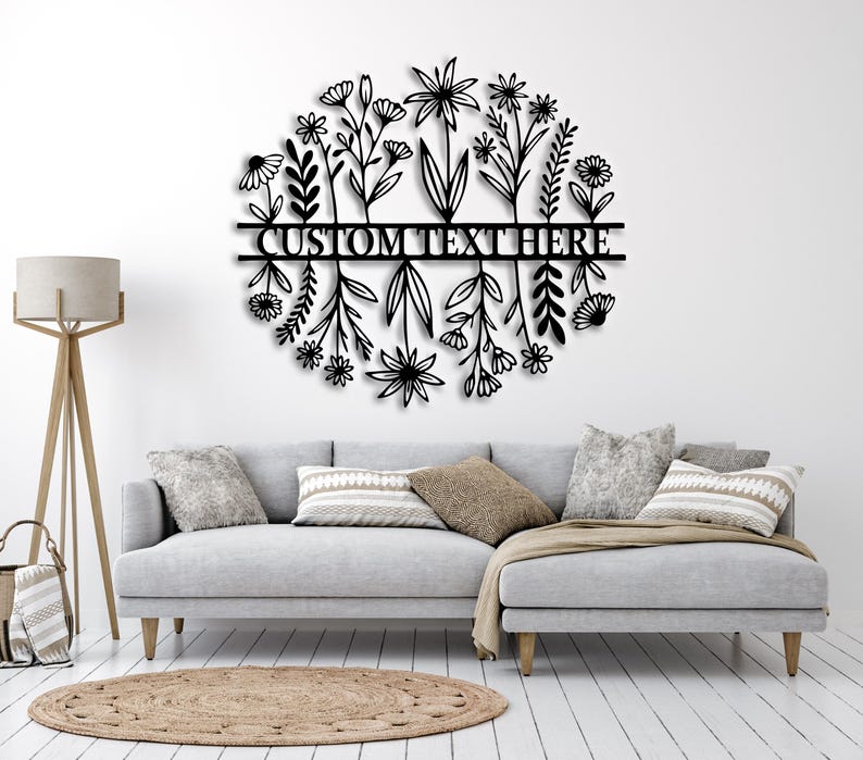 May include: Black metal floral wall art in a circular design. The artwork features stylized flowers and foliage around a horizontal bar with the text "CUSTOM TEXT HERE". It is mounted on a white wall above a grey sofa.