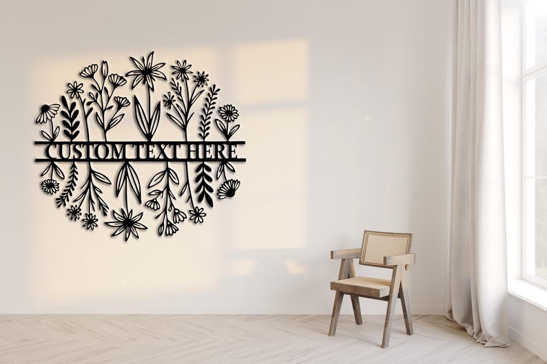 May include: Black metal floral wall art with the text "CUSTOM TEXT HERE" in a circular design. The artwork features various stylized flowers and foliage. The piece is mounted on a white wall, with a wooden chair and window in the background.