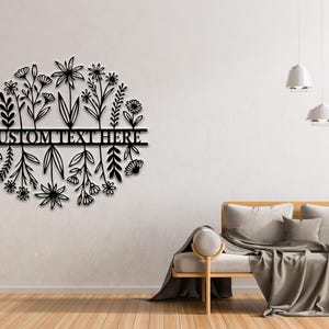 May include: Black metal floral wall art with the text "CUSTOM TEXT HERE" in a horizontal bar. The design features stylized flowers and foliage in a circular shape. The art is mounted on a white wall.