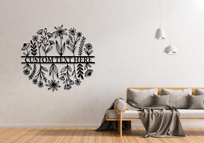 May include: Black metal floral wall art with the text "CUSTOM TEXT HERE" in a horizontal bar. The design features stylized flowers and foliage in a circular shape. The art is mounted on a white wall.