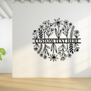 May include: Black metal floral wall art with the text "CUSTOM TEXT HERE" in a circular design. The artwork features various stylized flowers and leaves. The piece is mounted on a white wall in a bright room.