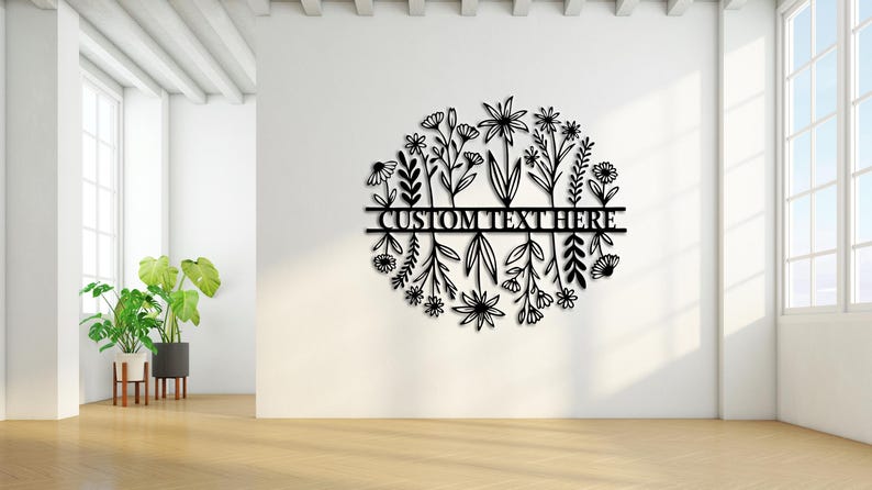 May include: Black metal floral wall art with the text "CUSTOM TEXT HERE" in a circular design. The artwork features various stylized flowers and leaves. The piece is mounted on a white wall in a bright room.