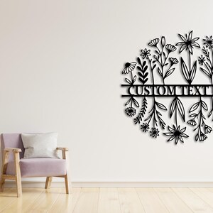 May include: Black metal floral wall art with the text "CUSTOM TEXT HERE" in a circular design. The art features various flower and leaf silhouettes. The wall art is mounted on a white wall.