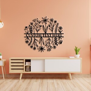 May include: Black metal floral wall art with a circular design featuring various flower and leaf silhouettes. The words "CUSTOM TEXT HERE" are displayed across the centre. The artwork is mounted on a peach-coloured wall above a wooden cabinet.