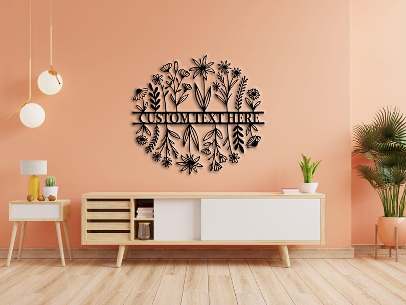 May include: Black metal floral wall art with a circular design featuring various flower and leaf silhouettes. The words "CUSTOM TEXT HERE" are displayed across the centre. The artwork is mounted on a peach-coloured wall above a wooden cabinet.