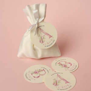 Peut inclure: Un sac de faveur en satin blanc noué avec un ruban, accompagné de trois étiquettes circulaires. Les étiquettes présentent un motif floral avec la lettre "A", le nom "Aurora" et la date "25 Maggio 2026".