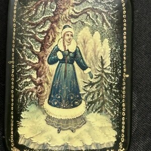 May include: A rectangular, black lacquered box featuring a hand-painted winter scene. The artwork shows a woman in a blue coat with white fur trim in a snowy forest. Gold trim borders the box and the image.