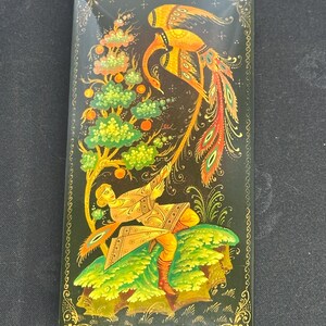 May include: Rectangular black lacquer box with a detailed painting of a golden bird with a long, colourful tail, a figure, and a tree with orange fruit. The box has a gold border and intricate floral designs.