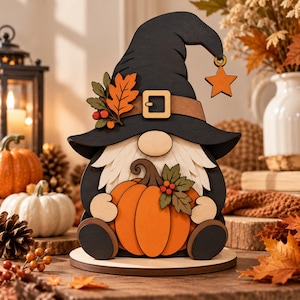 Op de afbeelding: Een houten kabouterfiguur met een zwarte puntmuts met een beige riem, een gesp en een oranje sterretje. De kabouter heeft een witte baard, houdt een grote oranje pompoen vast en is versierd met herfstbladeren en rode bessen op de muts en pompoen.