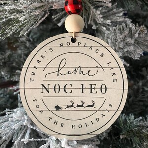 Op de afbeelding: Een ronde, crèmekleurige kerstversiering met de zwarte tekst "There's No Place Like Home For The Holidays". Het heeft een houten kraal en een rood-zwarte buffelruitkraal. De versiering hangt aan een besneeuwde kerstboom.