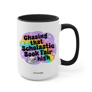 May include: White ceramic mug with a black rim and handle. The mug features a rainbow design with the text "Chasing that Scholastic Book Fair high" and a stack of books.