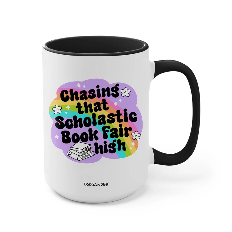 May include: White ceramic mug with a black rim and handle. The mug features a rainbow design with the text "Chasing that Scholastic Book Fair high" and a stack of books.