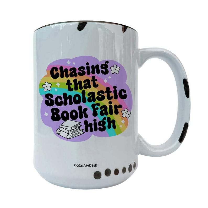 May include: White ceramic mug with a black handle and black speckles. The mug has a rainbow design with the text "Chasing that Scholastic Book Fair high" and a stack of books.