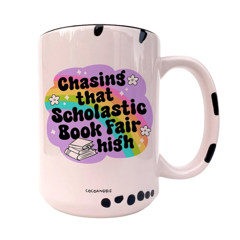 May include: White ceramic mug with a black rim and handle. The mug has a black and white polka dot pattern around the bottom. The mug features a rainbow design with the text "Chasing that Scholastic Book Fair high" and a stack of books.