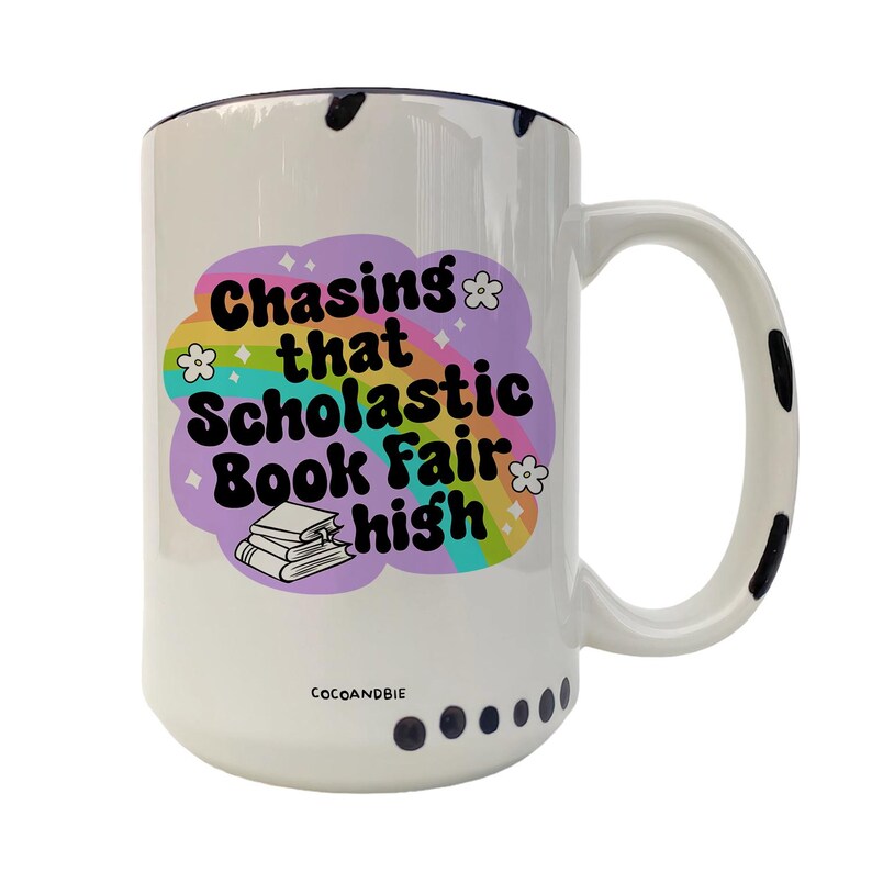 May include: White ceramic mug with a black rim and black polka dots around the bottom. The mug features a rainbow design with the text "Chasing that Scholastic Book Fair high".