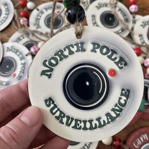 May include: A white ceramic ornament with the words "NORTH POLE SURVEILLANCE" in green lettering. The ornament features a black camera lens design with a red dot. The ornament is round and has a twine loop for hanging.