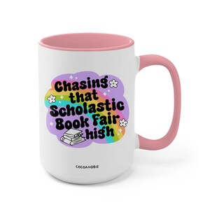 May include: White ceramic mug with a pink rim and handle. The mug features a rainbow design with the text "Chasing that Scholastic Book Fair high" and a stack of books.