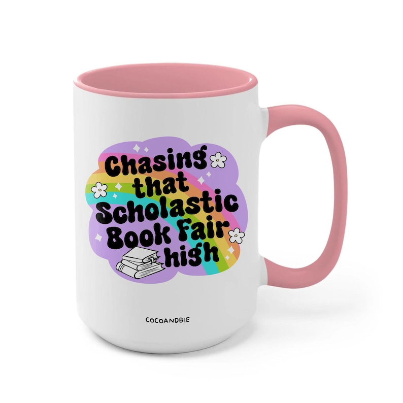 May include: White ceramic mug with a pink rim and handle. The mug features a rainbow design with the text "Chasing that Scholastic Book Fair high" and a stack of books.