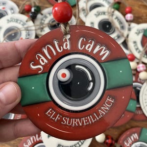 May include: A round Christmas ornament with a red and green design. The ornament features a camera lens graphic and the words "Santa Cam" and "Elf Surveillance" in white lettering. A red bead hangs from a twine loop.