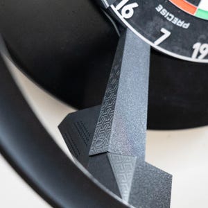 Könnte beinhalten: Nahaufnahme einer schwarzen und grauen Dartscheibe mit Ständer. Die Dartscheibe hat weiße Zahlen und das Wort "PRECISE" aufgedruckt. Der Ständer besteht aus strukturierten grauen und schwarzen Materialien mit geometrischen Mustern.