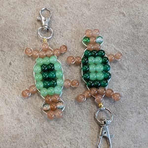 May include: Two beaded turtle keychains. Each turtle has a body made of green beads, with tan bead limbs and head. The keychains have silver-colored clasps.