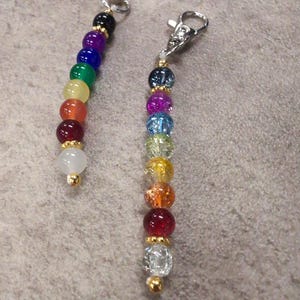 Puede incluir: Dos llaveros de cuentas de colores arcoíris con cierres plateados. Las cuentas son de diferentes tonos de rojo, naranja, amarillo, verde, azul, morado y blanco.