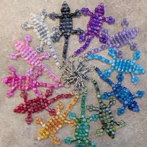 May include: A set of 12 beaded lizard keyrings in various colours, including pink, purple, blue, green, yellow, and black. The lizards are all made of small, round beads and have a silver key ring attached.
