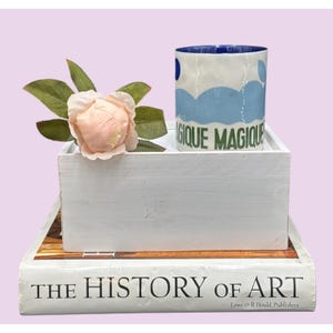 May include: A white ceramic mug with blue cloud designs and the words "MAGIQUE MAGIQUE" in green. The mug sits on a white wooden box, next to a pink flower and green leaves. The box rests on a book titled "THE HISTORY OF ART".