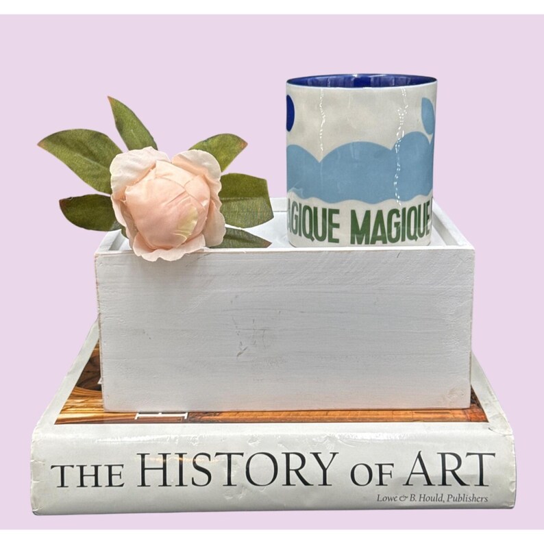 May include: A white ceramic mug with blue cloud designs and the words "MAGIQUE MAGIQUE" in green. The mug sits on a white wooden box, next to a pink flower and green leaves. The box rests on a book titled "THE HISTORY OF ART".