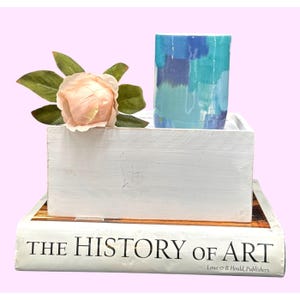 May include: A white wooden box with a light pink flower and green leaves, topped with a blue and teal painted ceramic cup. The box rests on a book titled "THE HISTORY OF ART".