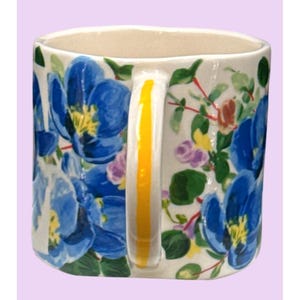 May include: A ceramic mug featuring a white base decorated with large blue floral patterns and yellow centers. The handle is white with a vertical yellow stripe. Green leaves and smaller purple and yellow flowers complete the design. The interior is cream-coloured.