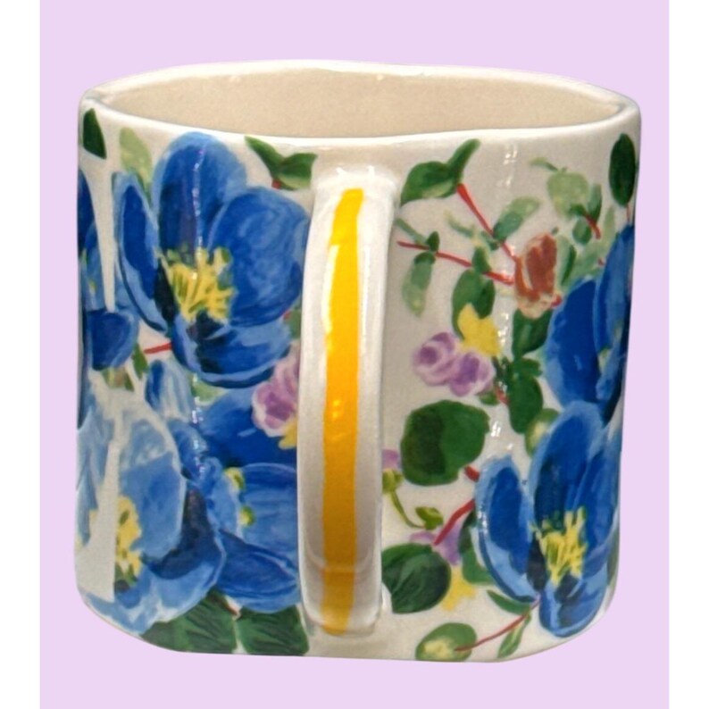 May include: A ceramic mug featuring a white base decorated with large blue floral patterns and yellow centers. The handle is white with a vertical yellow stripe. Green leaves and smaller purple and yellow flowers complete the design. The interior is cream-coloured.